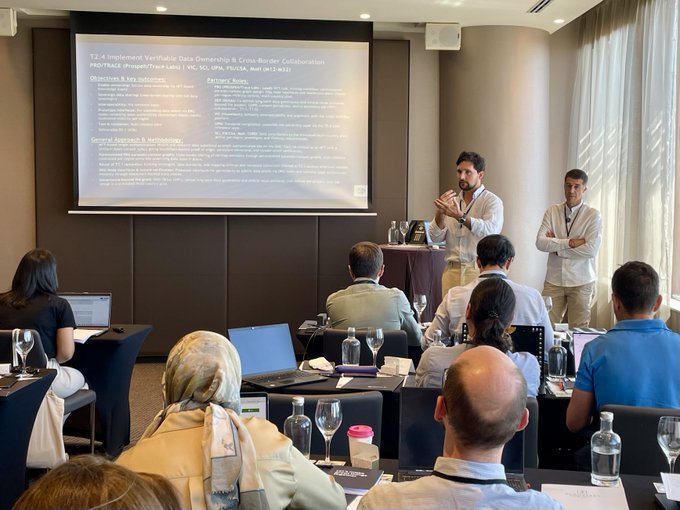 A photograph of a presentation in a conference room where two men stand before a slide titled "T3.4 Implement Verified in Data Ownership & Cross-Border Collaboration" while attendees watch.