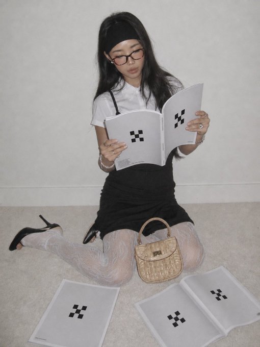 A young woman sits cross-legged on the floor in a black dress, white blouse, and patterned tights, reading books displaying the OKX logo, with a tan handbag and additional OKX-branded books arranged around her. The image ties to her announcement of becoming an OKX creator, with the branded materials symbolizing the partnership.