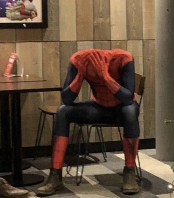 A person in a Spider-Man costume sits slumped over in a chair at a café, head in hands, wearing brown boots.