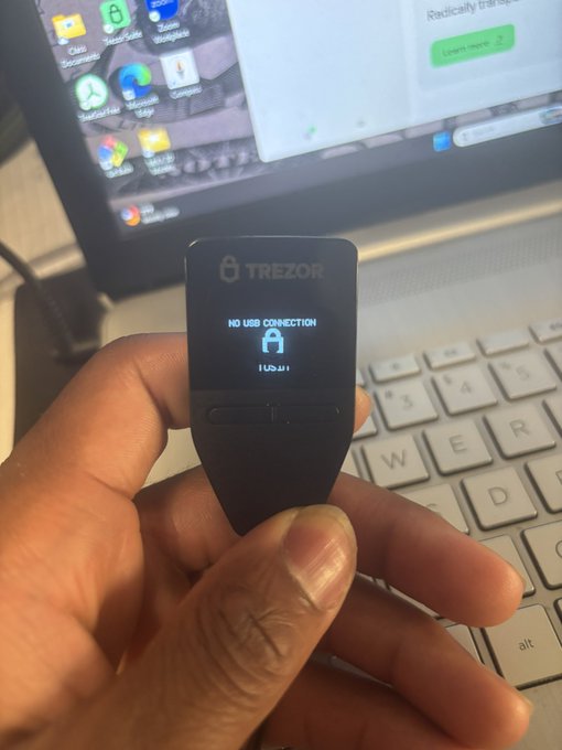 A hand holds a Trezor hardware wallet displaying a 'NO USB CONNECTION' lock screen, with a laptop showing app icons visible in the background.