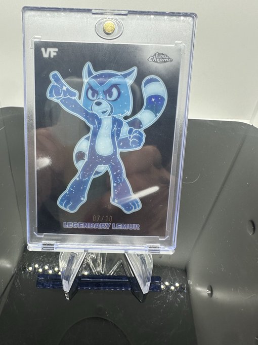 The image features a small, clear plastic card with a cartoon character design on it. The character appears to be a raccoon or a cat, standing upright and pointing its finger towards something out of frame. The background of the image is black, which contrasts with the white text that reads "VeeFriends" in the bottom right corner.