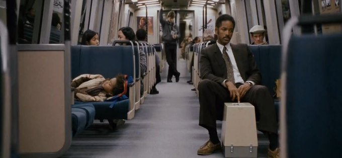 The image shows a man sitting on a train, wearing a suit and tie, with his head resting on his hand. The scene takes place inside a subway car where several people are seated or standing around him. One person appears to be sleeping while another is holding a suitcase. A third person can be seen walking down the aisle of the train.