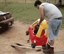 The image shows a man standing next to a red toy car, which appears to be a small car or truck for children. He is wearing blue jeans and a gray shirt. The scene takes place outdoors with a grassy area around them. In the background, there are two cars parked nearby - one being a pickup truck and another smaller vehicle. A caption below the image reads "GM #crypto #HappyFathersDay".