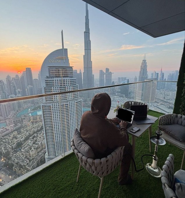 A woman wearing a hijab sits at a small table on a high-rise balcony overlooking the Dubai skyline at sunset, with the Burj Khalifa and the 'EMAAR' building visible in the distance.