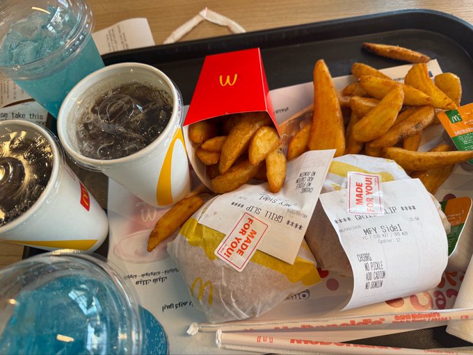 A McDonald's meal on a tray featuring fries, two burgers in 'MADE FOR YOU' wrappers (including one labeled 'Grilled Sl'), and McDonald's branded cups with blue drinks.