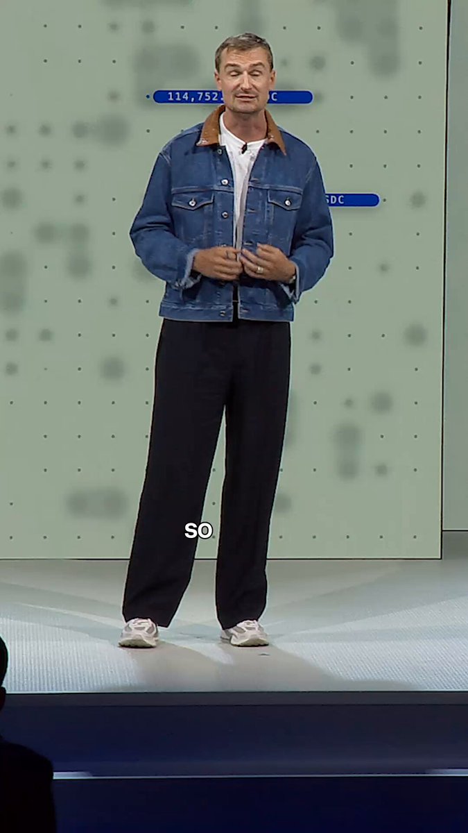 The image features an older man standing confidently on stage, wearing a blue denim jacket and black pants. He appears to be giving a speech or presentation, possibly at a conference or event. The background of the image shows a white wall with various signs hanging from it, suggesting that this is a professional setting where important information is being shared.