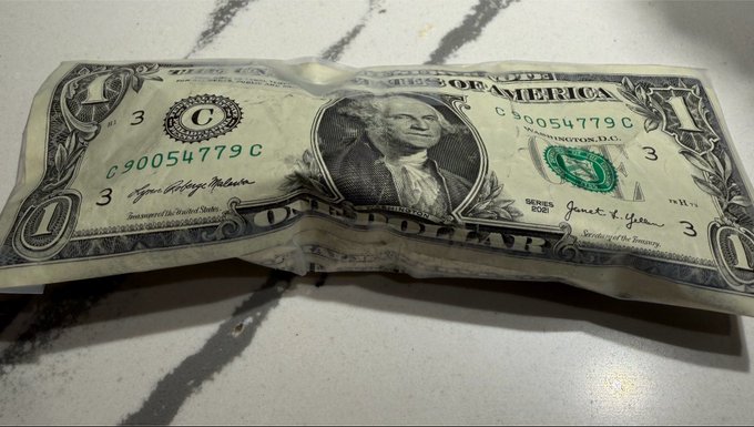 A crumpled one-dollar bill resting on a white surface with dark, marble-like streaks.