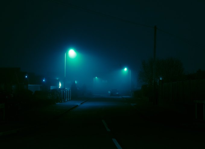 A moody, foggy nighttime street scene is illuminated by green-tinted streetlights casting an eerie glow on the deserted road.