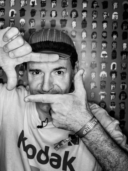 A black and white photograph of a man wearing a Kodak t-shirt and a flat cap, framing his eyes with his hands, standing in front of a backdrop covered in repeating illustrated face icons.