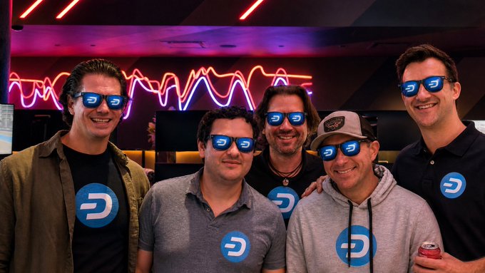 A group photo of six men at what appears to be a Dash cryptocurrency event, with digital Dash logos edited onto their faces and shirts, posed in front of a neon waveform light display.
