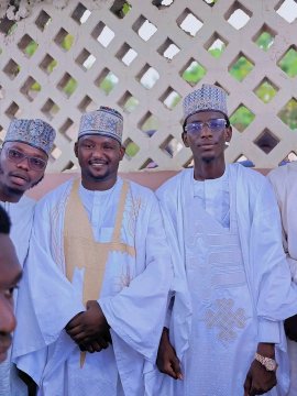 Three men in traditional white West African attire and embroidered caps smile together in front of a white lattice wall.