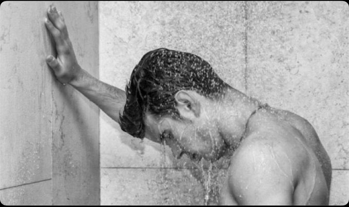 A black and white photograph shows a person leaning against a tiled wall while water pours over their head from above, creating a striking, artistic composition.