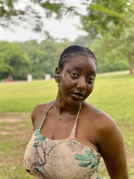 A portrait of a young woman with slicked-back braided hair wearing a floral print halter top, standing in an outdoor park setting with green trees and grass in the background.