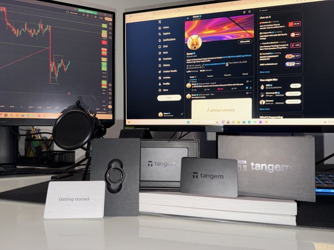 A desk setup featuring Tangem crypto hardware wallets displayed on a white platform, with two monitors in the background showing trading charts and X (Twitter) social media feeds.