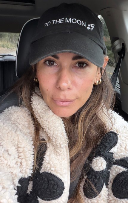 A woman wearing a black baseball cap with the text 'TO THE MOON' and a rocket logo, along with a black and white sherpa fleece jacket, taking a selfie inside a car.