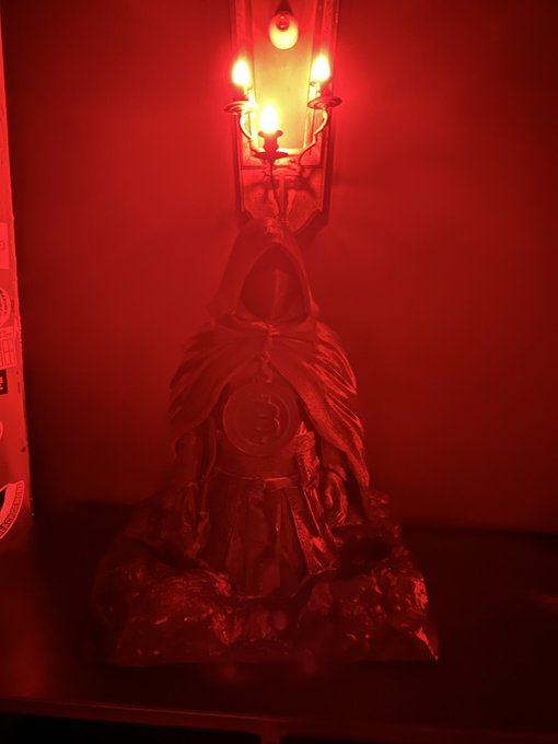 A hooded figurine resembling Doctor Strange is illuminated by a candelabra in a dark, red-lit setting, evoking an ominous yet magical atmosphere. This relates to the post "He's Home" by referencing the mystical, mystical homecoming vibe associated with the character.