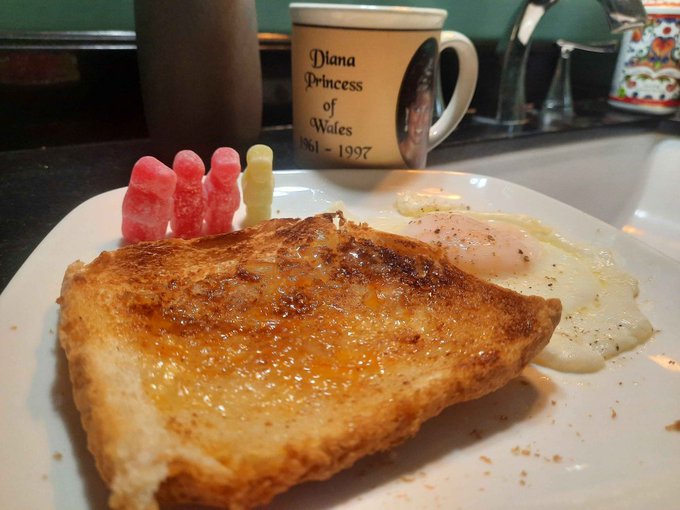 A plate of golden-brown French toast served alongside a sunny-side-up fried egg and pink/white gummy candies, with a commemorative 'Diana Princess of Wales 1961-1997' mug visible in the background.