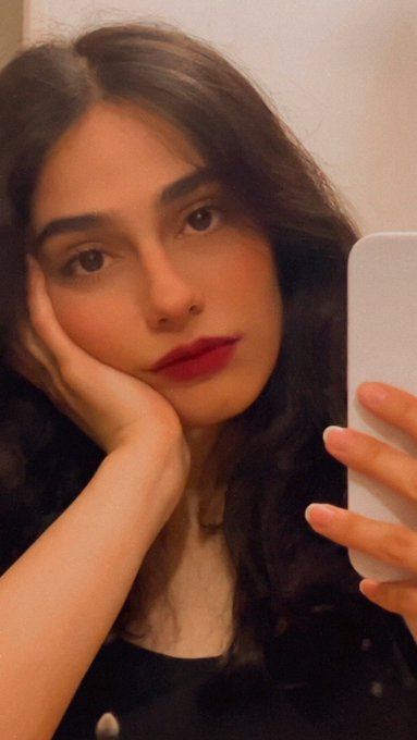 A selfie of a young woman with dark wavy hair and red lipstick, resting her cheek on her hand while holding a smartphone.