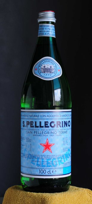 A green glass bottle of S. Pellegrino sparkling natural mineral water (100 cL) sitting on a yellow towel against a dark background.