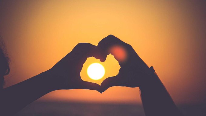 A silhouette photograph of two hands forming a heart shape, framing the bright orange sun setting over the ocean.