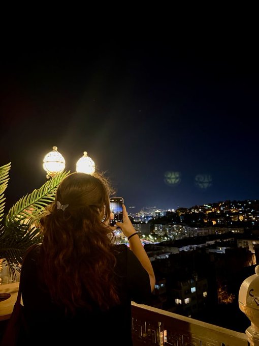 A woman with long hair takes a photo of a city skyline at night with her smartphone from a balcony illuminated by a lamp.