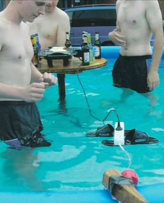 Three shirtless men stand in a swimming pool around a small wooden table holding beer bottles, with electrical devices, sandals, and a phone floating in the water.