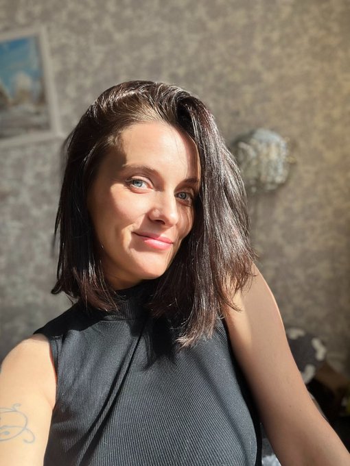 A close-up selfie of a smiling young woman with long dark hair and blue eyes, wearing a black sleeveless top, with bright sunlight illuminating her face.