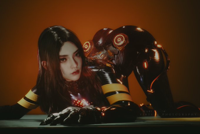 A cosplayer poses in a sleek black and yellow armored suit with glowing orange accents, embodying the character Tachy from Stellar Blade. The watermark "Marc Herite Photography" is visible in the bottom right corner.