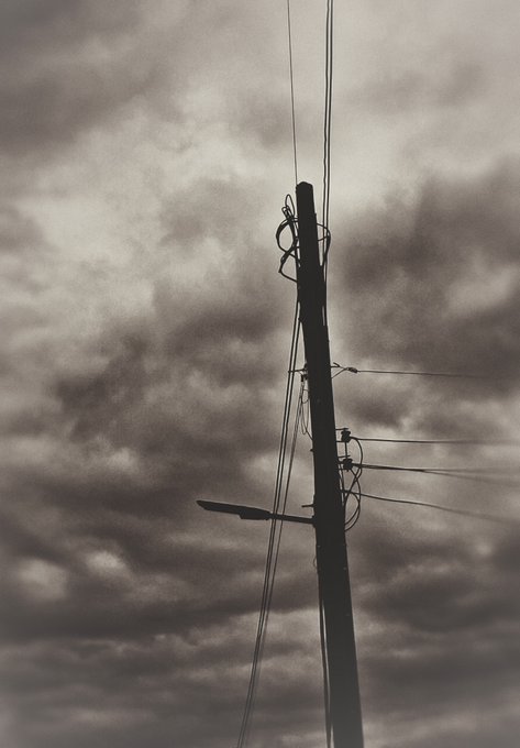 A black-and-white silhouette of a utility pole with tangled wires and a street lamp, set against a dramatic, cloudy sky.