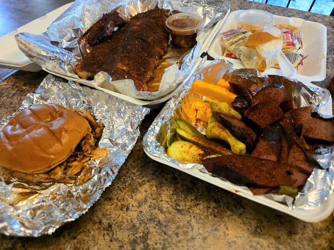 A spread of mouthwatering BBQ takeout, including pulled pork and brisket sandwiches, ribs, sausage links, and classic sides like pickles, cheese cubes, and bread rolls, all served in foil-lined trays on a granite countertop — fitting perfectly with the caption promoting a hearty "best BBQ in the world" giveaway in exchange for SOL wallets.