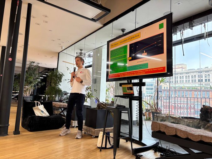The image shows a man standing behind a large screen, giving a presentation or speech. The room has a black and white color scheme with wooden floors and walls. There are two potted plants placed near the center of the room, one on each side. A chair is also visible in the background. The post includes text that reads "Thank you @jdetychey for speaking on $ETH issuance at yesterday'S All Protocol Devs Happy Hour! And thank you to @SeiNetwork for hosting our group again for these discussions."
