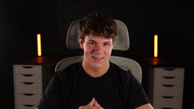 A young man with curly brown hair smiles at the camera while seated in a modern office chair, framed by two warm orange light bars on a dark background, with his hands clasped together. The setup, with its sleek lighting and professional vibe, suggests he's a content creator gearing up for a new video discussing the numerology behind $TROLL's potential rise.