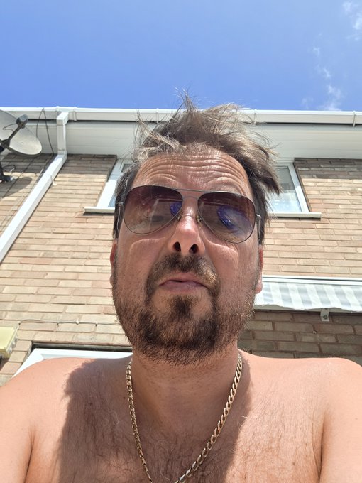 Low-angle selfie of a shirtless, bearded man wearing gold-rimmed aviator sunglasses and a gold chain, photographed against a brick house and blue sky.