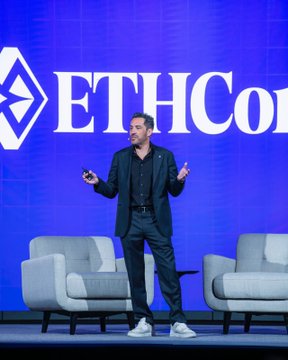 The image features a man standing confidently at a podium, wearing a suit and tie, with his hands outstretched as if he's giving a speech or presentation. The backdrop of the scene is a large blue screen displaying the name "ETHCON" in white letters. There are two chairs visible behind the man, one on each side of him.