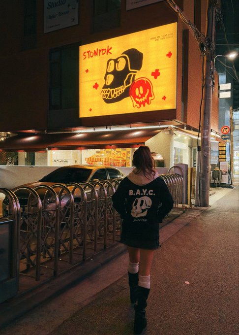A person wearing a black "B.A.Y.C." sweatshirt and skirt stands on a nighttime city street, gazing up at a large illuminated sign reading "STORYBOX" featuring a stylized skull design. The scene captures the bold streetwear aesthetic of the BAYC (Bored Ape Yacht Club) collection against the urban Seoul backdrop, tying together the neon-lit storefront with the iconic ape-themed apparel.