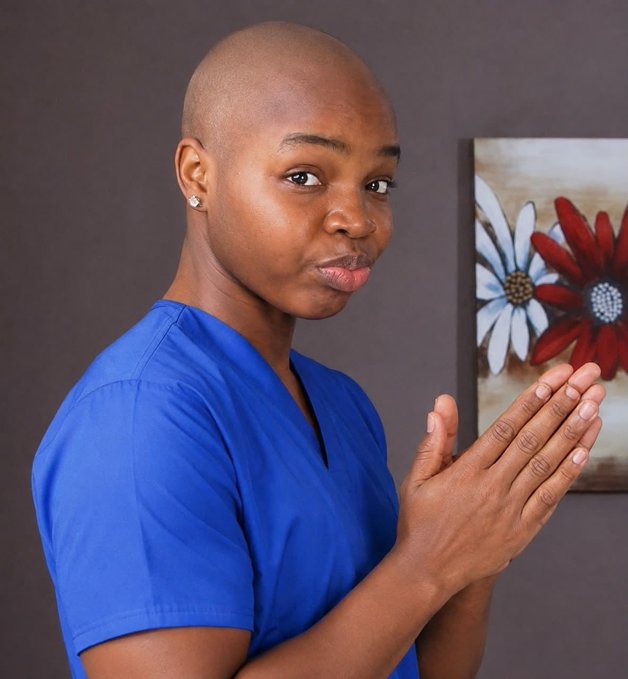 A portrait of a bald person wearing blue medical scrubs and stud earrings, posing with clasped hands in front of a wall with a floral painting.