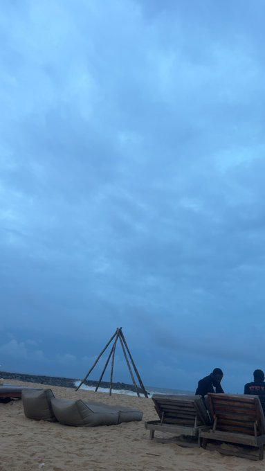 Under an overcast, twilight sky, two people sit on a wooden bench on a sandy beach beside a pile of beanbag chairs and a wooden tripod structure.