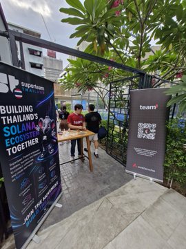 An outdoor event booth setup with banners reading 'Building Thailand's Solana Ecosystem Together' and 'Team Superteam Thailand', where two people are working at a table.