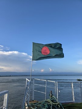 The flag of Bangladesh, featuring a green field with a red circle, flies over a railing with the sea and a cloudy sky in the background.