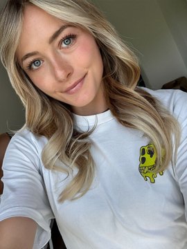 The image features a young woman wearing a white t-shirt with a cartoon skull design on the front. She has blonde hair, which complements her outfit. The photo captures her smiling face as she poses for the camera. The caption below the image reads "Mondays are for Mutants Now.. show me your Merch HMM". This suggests that the woman is promoting a brand or merchandise related to the cartoon skull design on her t-shirt, possibly indicating that she is a fan of the character or wants to share her enthusiasm with others.