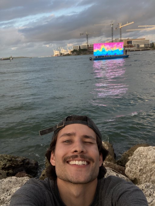 A man takes a selfie by the water, smiling with one eye closed, while a boat with a large digital screen glows in the background.