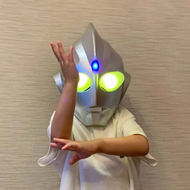 A child wearing an Ultraman mask with glowing yellow eyes and a blue forehead light, posing with arms crossed in front of a beige textured wall.