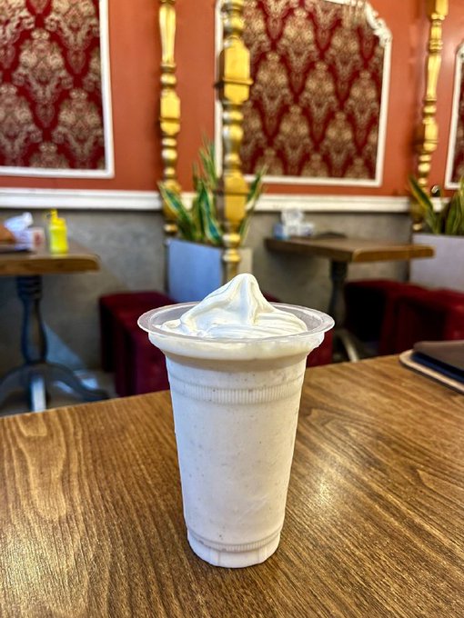 A whipped cream-topped milkshake or float in a clear plastic cup sits on a wooden table in a restaurant with red patterned wall panels and green plants in the background.