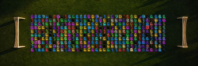 An aerial view shows dozens of brightly colored luxury cars arranged in a precise rectangular grid on a grassy field, flanked by two helicopter landing pads.