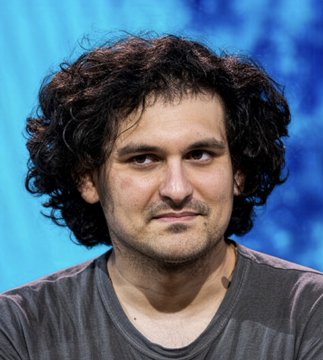 A photo of a man with curly dark hair wearing a gray t-shirt against a blue background.