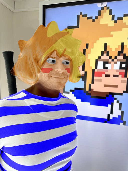 A person wearing an orange and yellow wig and a blue and white striped shirt stands in front of a pixel art display of a similar character with blonde hair.