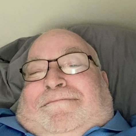 A close-up selfie shows a man with glasses, a gray beard, and a blue shirt lying down with his eyes closed, resting his head on a gray pillow. The image accompanies his announcement of revealing his face to his social media followers now that the algorithm has returned and his friends are present.