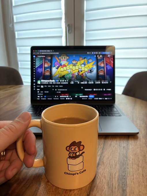 A hand holds a beige mug labeled 'Chimpi's Café' with a cartoon chimp logo, in front of a laptop displaying a website with 'CHIMPERS' branding.