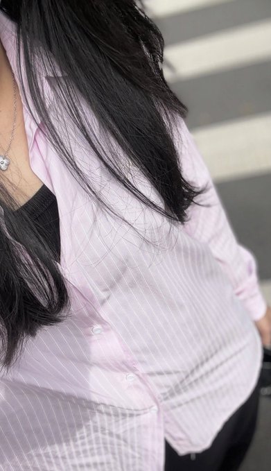 A person with long black hair wearing a light pink striped button-up shirt over a black top is photographed from the chest down, wearing a silver necklace.