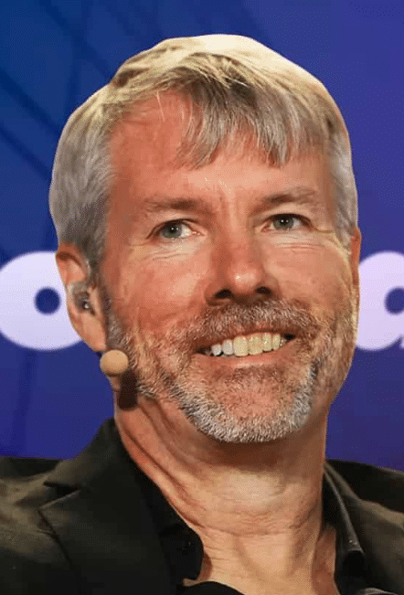 A smiling man with gray hair and a beard, wearing an earpiece microphone and a black blazer, speaks at an event (appearing to be MicroStrategy-related based on the partial logo visible in the background). The image accompanies news of Michael Saylor's Strategy achieving a $2.5 billion unrealized gain, a dramatic turnaround from the $8.1 billion unrealized loss reported just two weeks prior.